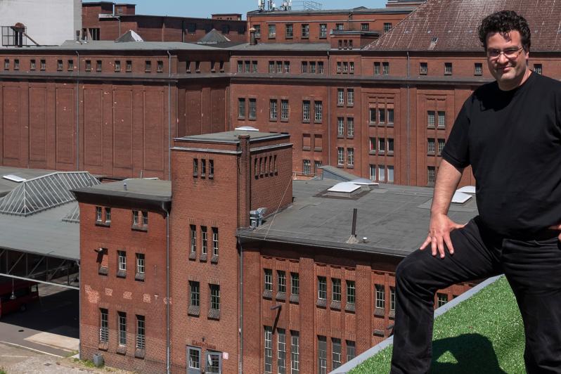 Portrait von Carsten Bredow auf dem Dach eines der Gebäude auf dem Gelände der Malzfabrik. Unter seinen Füßen ist ein Stück grüner Teppich und er hat ein Bein auf das Dachsims gestellt. Im Hintergrund ist das Hauptgebäude mit den vier Darren.