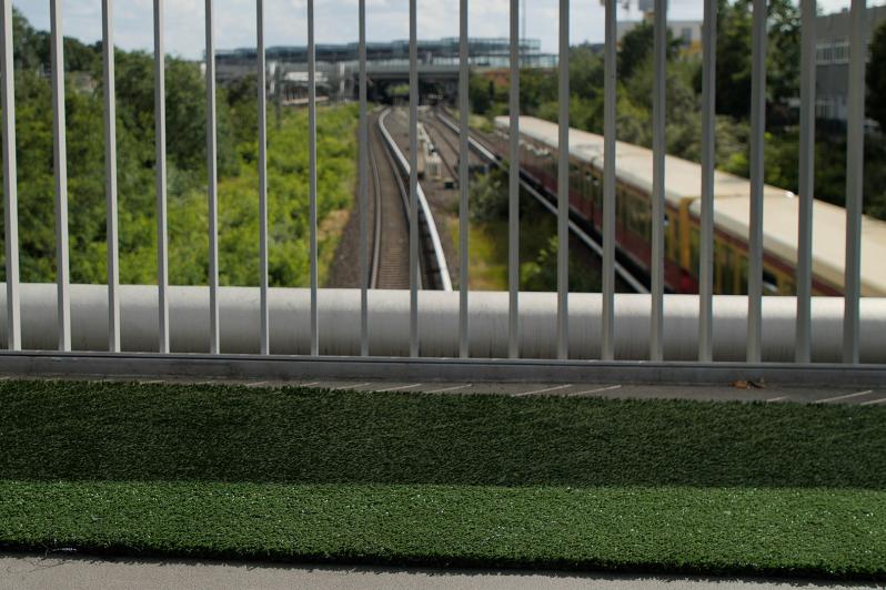 Grüner Rollrasen rollt üer eine Brücke unter der ein Zug fährt.